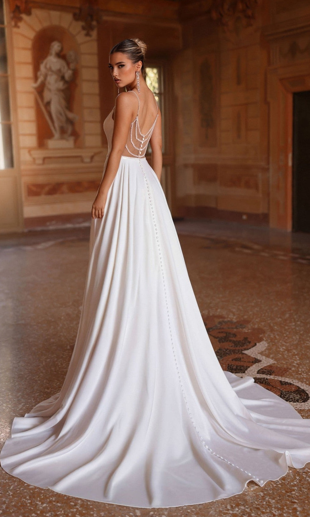 A woman wearing the Alyce 7099 long white dress with beaded spaghetti straps and an open back stands in an ornate room with classical decor and a marble floor.